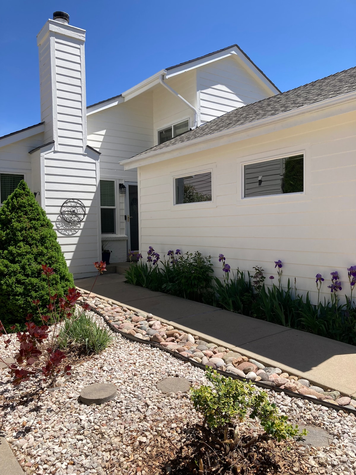 A welcoming exterior showcases a white house with a sloped roof and a prominent chimney. Colorful flowers and vibrant shrubs line the pathway, leading to the entrance. Natural stones and mulch create a landscaped bed, enhancing the overall appeal of the home.