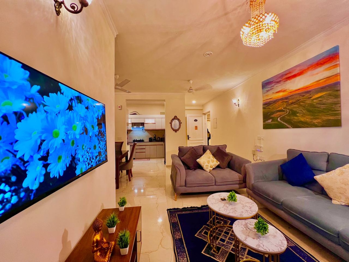 A spacious living area features a comfortable gray sofa and two marble-topped coffee tables. A large flat-screen TV displays vibrant blue flowers on the wall. Modern artwork adorns the opposite wall, and soft lighting enhances the inviting ambiance in the room.