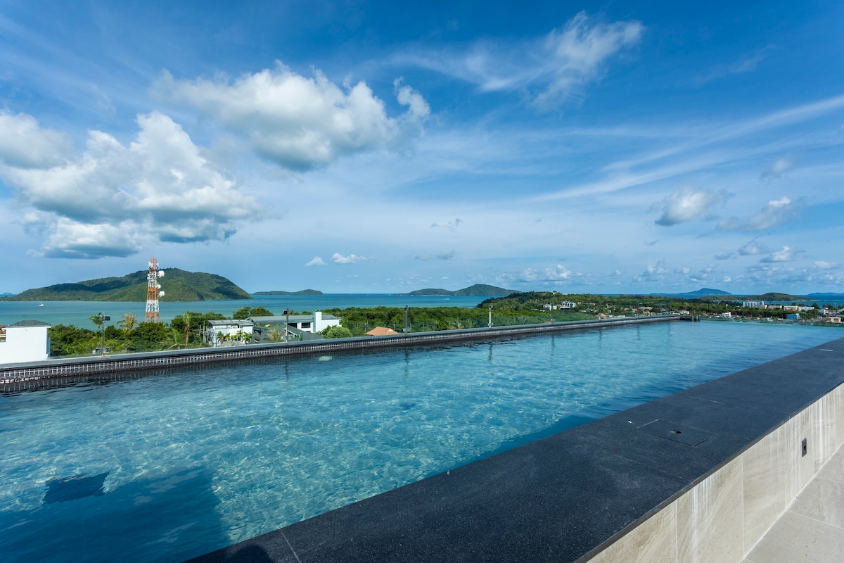 An infinity pool stretches along the edge of a rooftop, offering panoramic views of the ocean and surrounding islands. The clear water reflects the bright blue sky and fluffy white clouds, creating a serene environment for relaxation.
