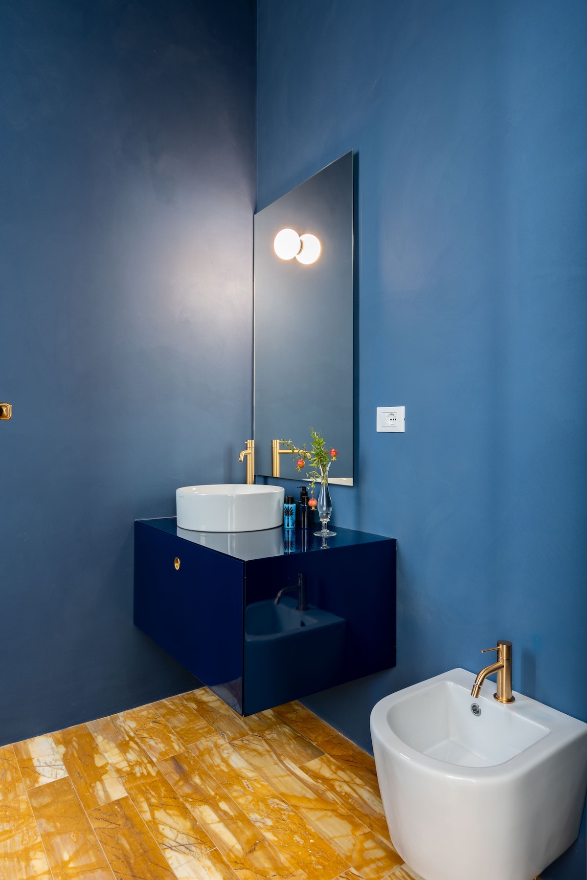A modern bathroom features a stylish blue wall with a large mirror above a sleek vanity. A round, white basin is set on a dark blue cabinet, while a bidet sits alongside. Soft yellow tiles provide a warm contrast to the cool blue tones.