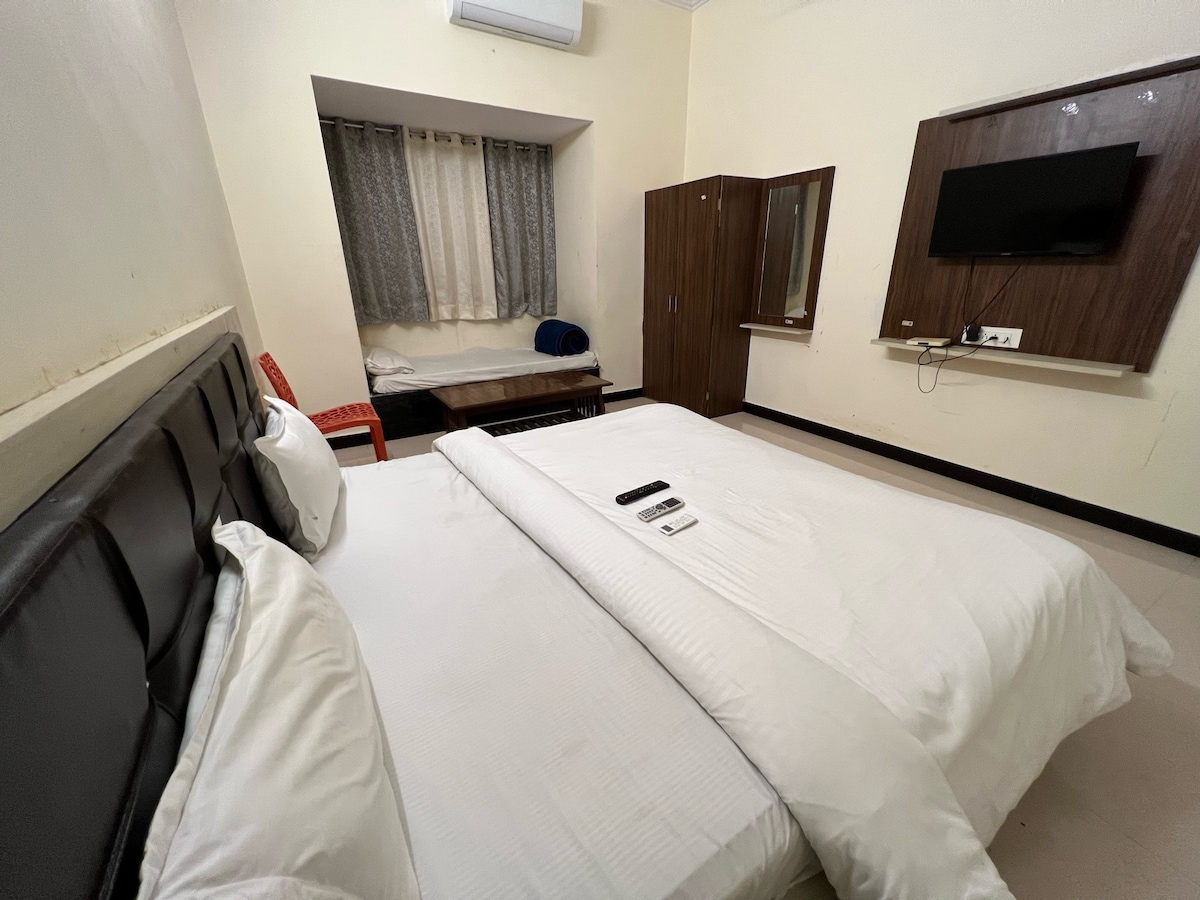 A spacious room features two large beds with white linens, positioned against a dark headboard. A window with light curtains allows natural light to enter, while a wooden cabinet and a television are clearly visible. A red chair and a bench are also included in the room.