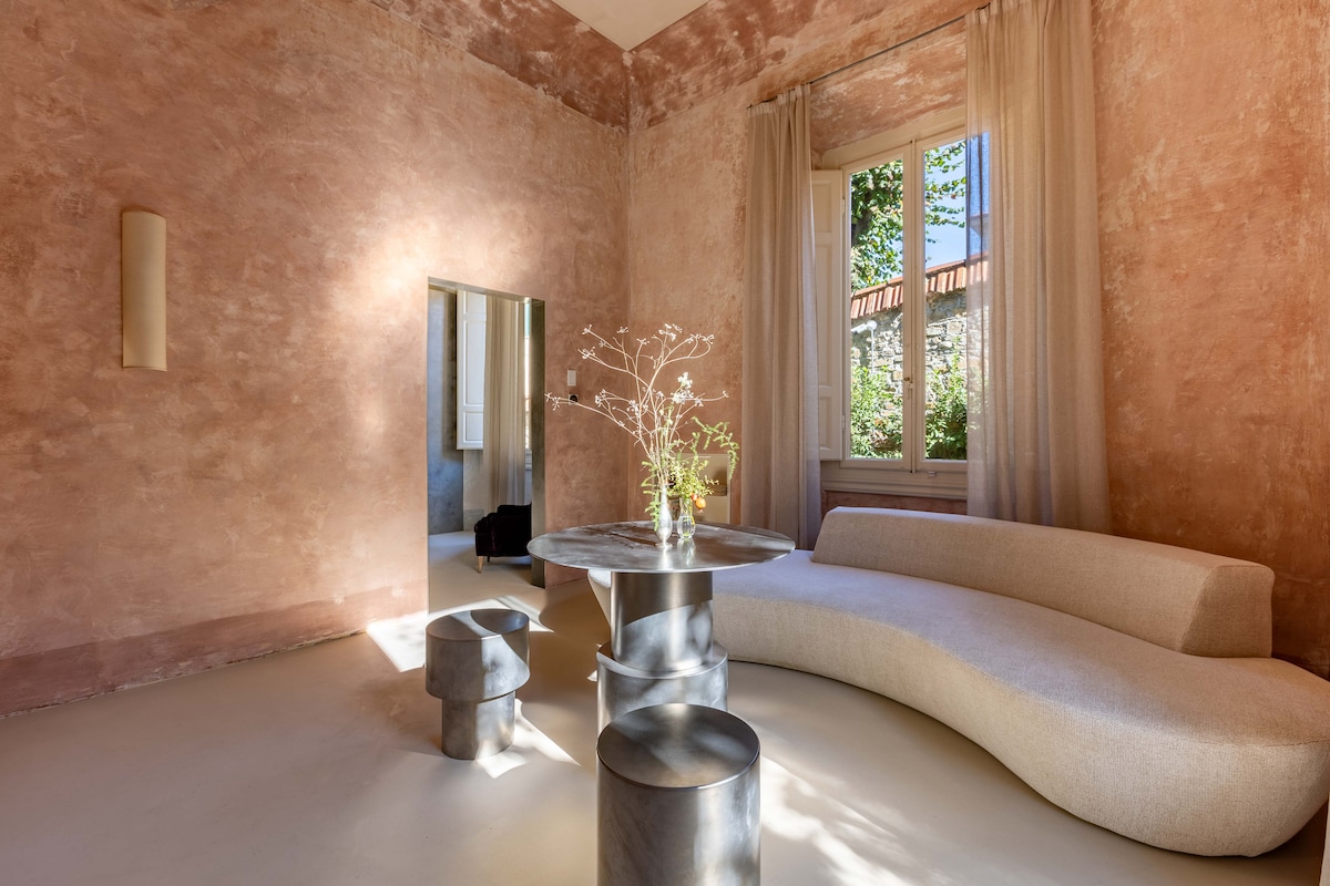 A sunlit living space features a curved sofa and a round table. A tall window allows natural light to fill the room, highlighting the textured pink walls. A mirror reflects part of the room, and a minimalist vase holds delicate branches.