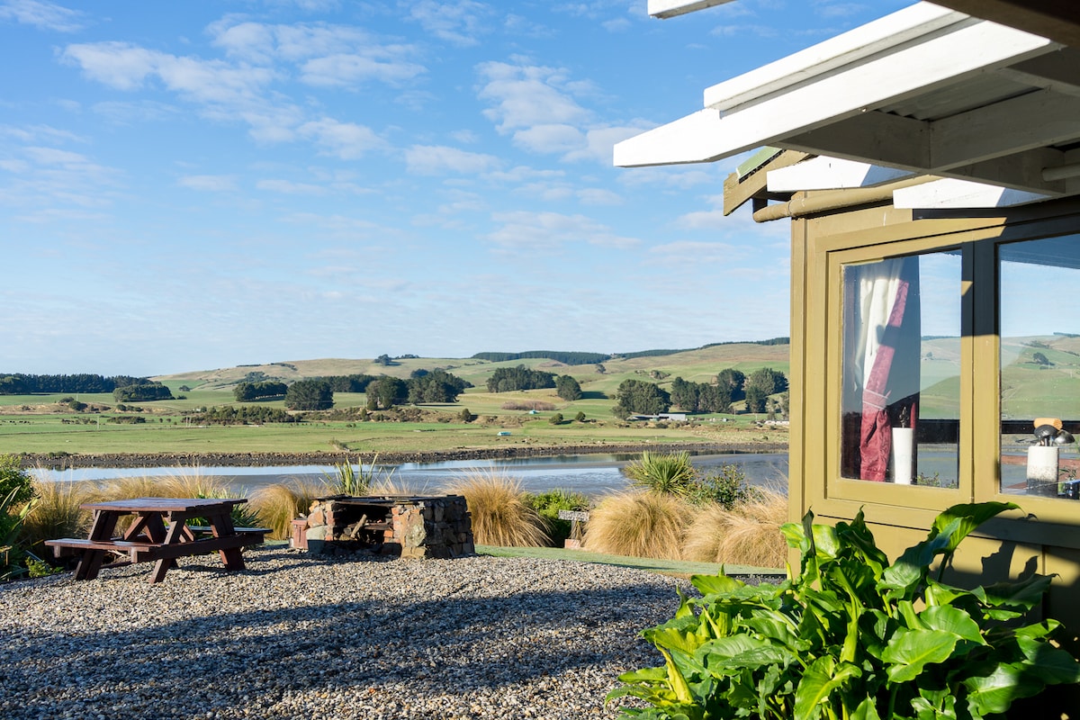 The exterior of the house is framed by lush greenery and gravel pathways. A picnic table is visible in the foreground, with a picturesque landscape of rolling hills and an estuary in the background, complemented by a clear blue sky.
