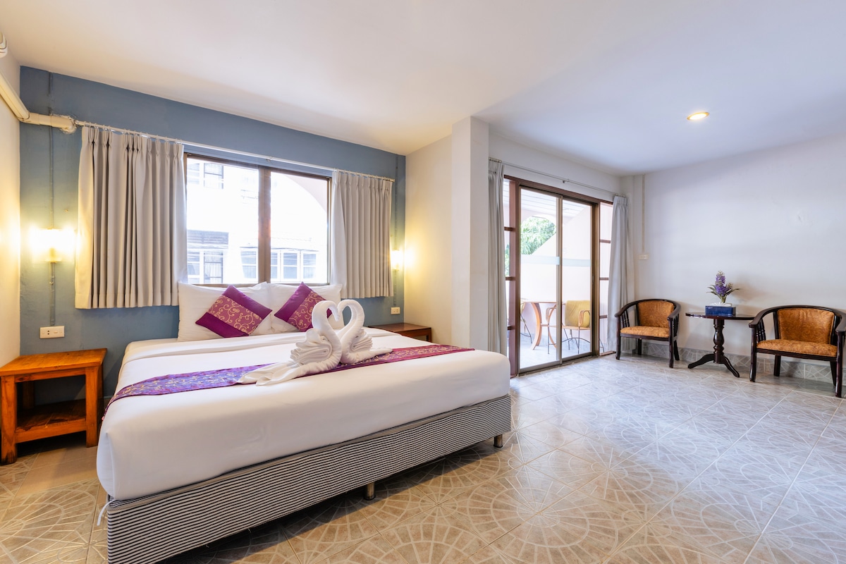 A spacious room features a large bed adorned with decorative pillows. A towel sculpture is positioned at the foot of the bed. Natural light enters through wooden-framed windows, which open to a small outdoor seating area. A wooden table and two chairs are visible.