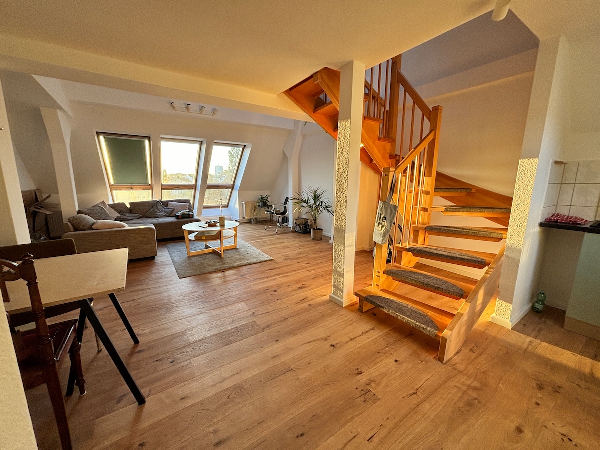 A spacious living area features large triangular windows that draw in natural light. A comfortable sofa is visible, accompanied by a coffee table. A staircase with wooden steps leads to an upper level, while a small dining table is placed in a corner.