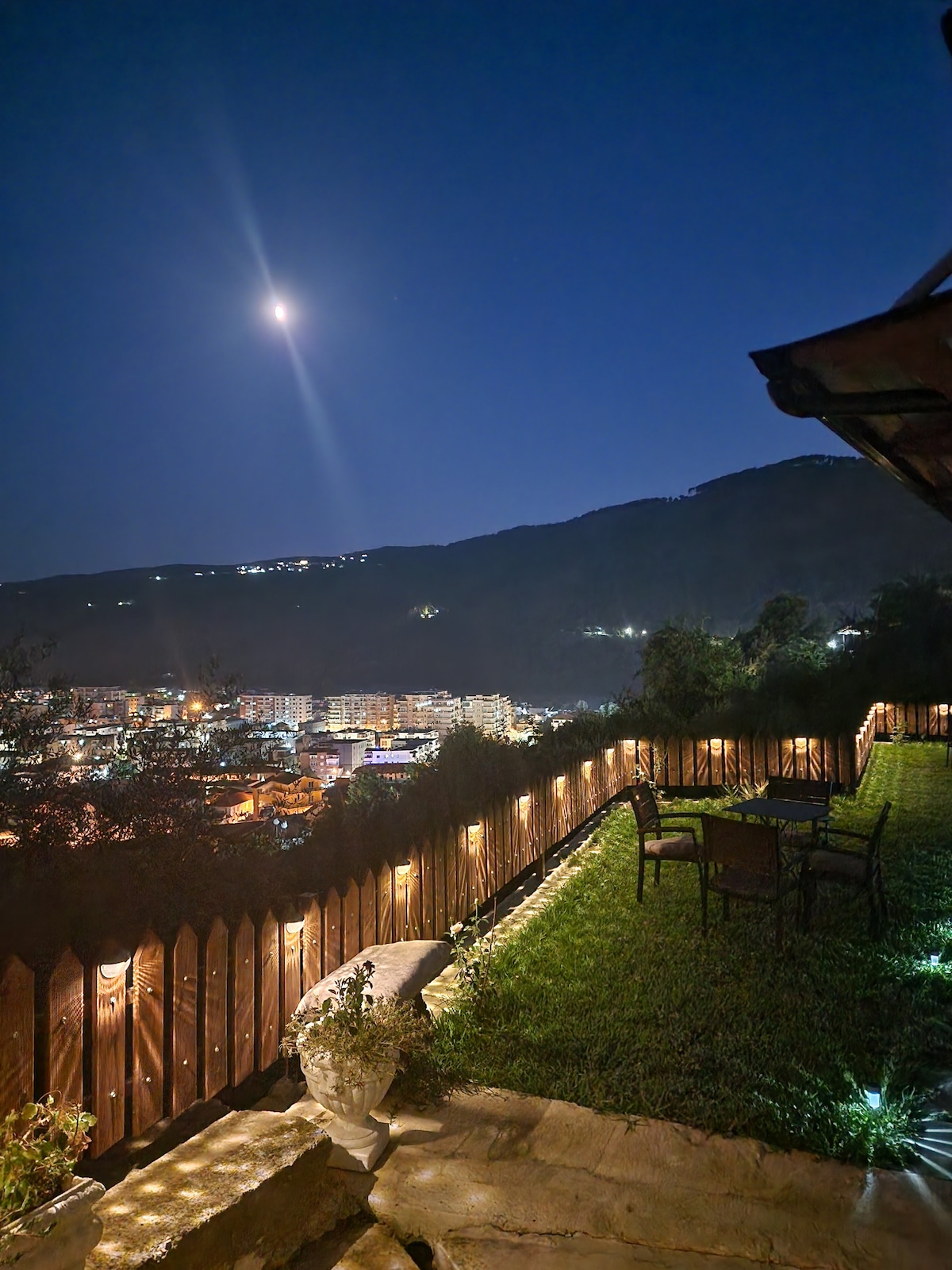 A scenic outdoor space is illuminated by soft lighting at night, featuring a manicured lawn bordered by a wooden fence. A small table with chairs offers a relaxing area to appreciate the views of the surrounding mountains and city lights under a clear moonlit sky.