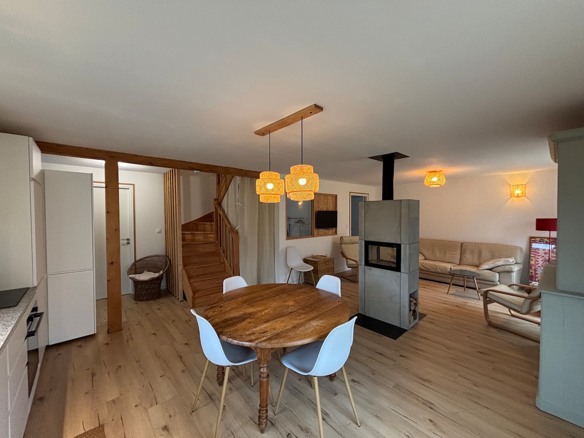 A spacious living area features a round wooden dining table surrounded by white chairs. Two stylish light fixtures hang above the table. A cozy seating arrangement is situated next to a modern fireplace, with a staircase leading to the upper level. Natural light brightens the space.