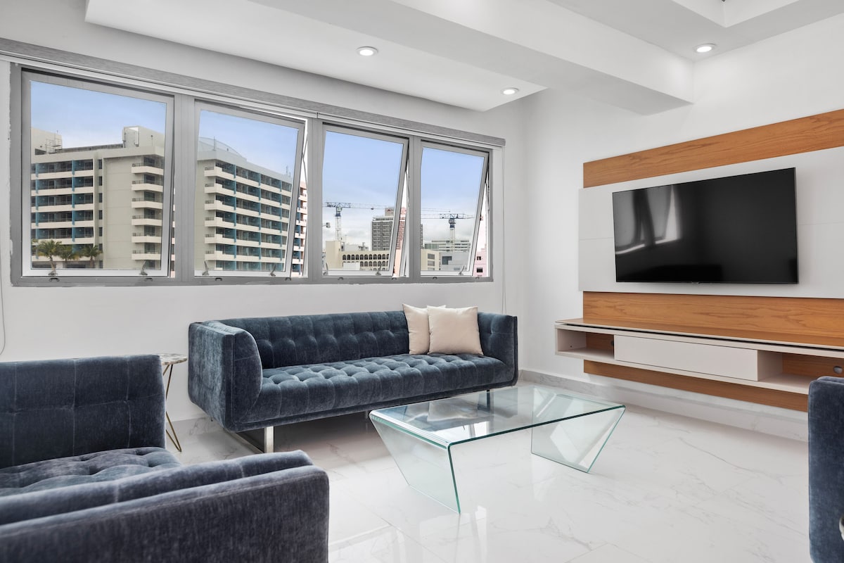 A bright living area features modern furnishings, including a plush blue sofa and armchairs. A glass coffee table sits centrally, while large windows allow natural light to fill the room. A wooden media console holds a flat-screen television, enhancing the contemporary design.