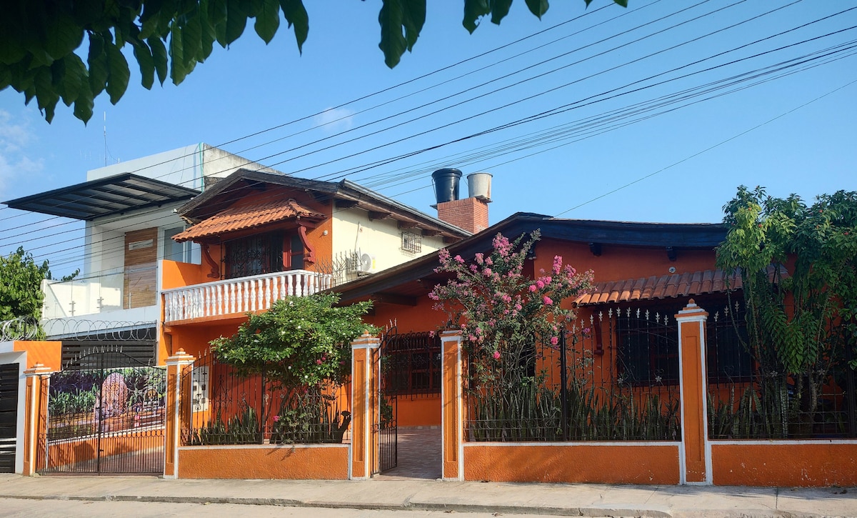 The exterior of El Jaguar Casa Hotel is showcased with vibrant orange walls and lush greenery. A balcony is visible on the upper level, and decorative ironwork frames the welcoming entryway. The surrounding area features well-maintained plants and a clear blue sky overhead.