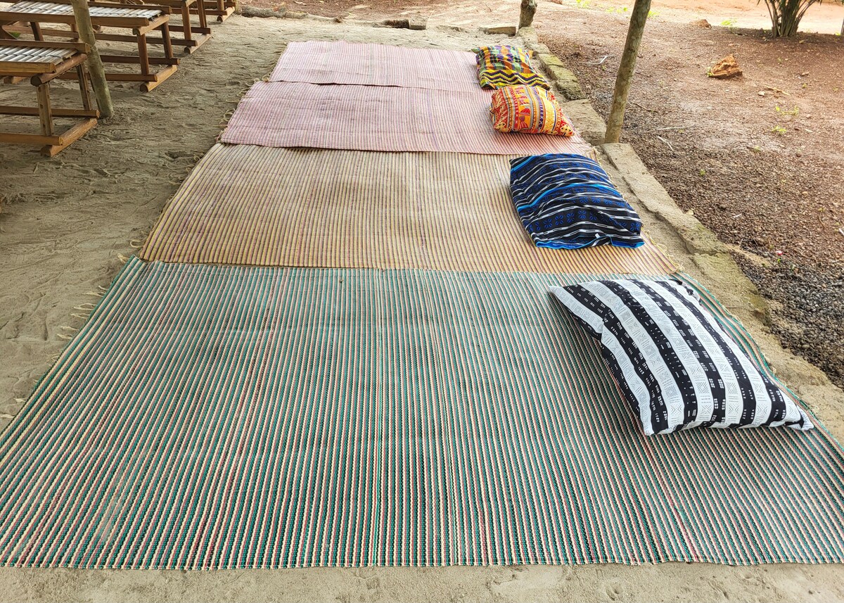 A series of woven mats are arranged on a flat, sandy surface, providing a comfortable setting for relaxation. Colorful pillows are placed alongside the mats, adding soft resting options for guests. The area is surrounded by natural elements, enhancing the serene environment.
