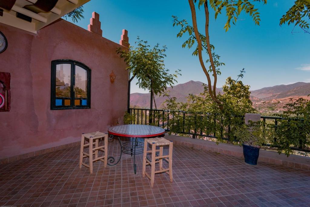 An outdoor terrace is shown, featuring a round table surrounded by two wooden stools. Lush greenery from surrounding trees and mountains is visible in the background, enhancing the serene atmosphere. The space offers clear views of the landscape beyond.