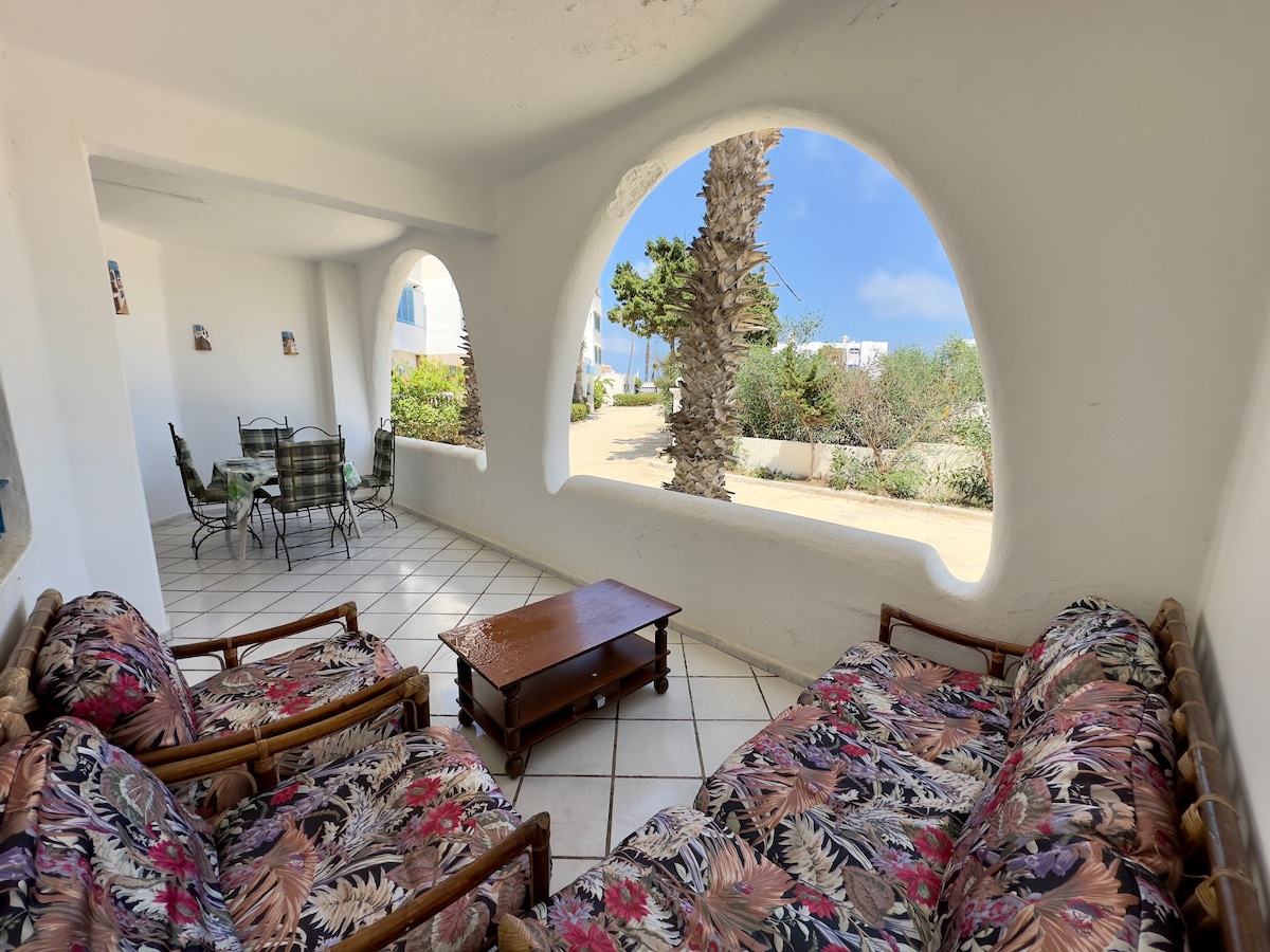 A spacious outdoor seating area is featured, showcasing patterned sofas and a wooden coffee table. Arched windows provide views of the lush greenery outside, with sun-drenched tiles beneath. The space promotes relaxation and connection with nature in a serene environment.