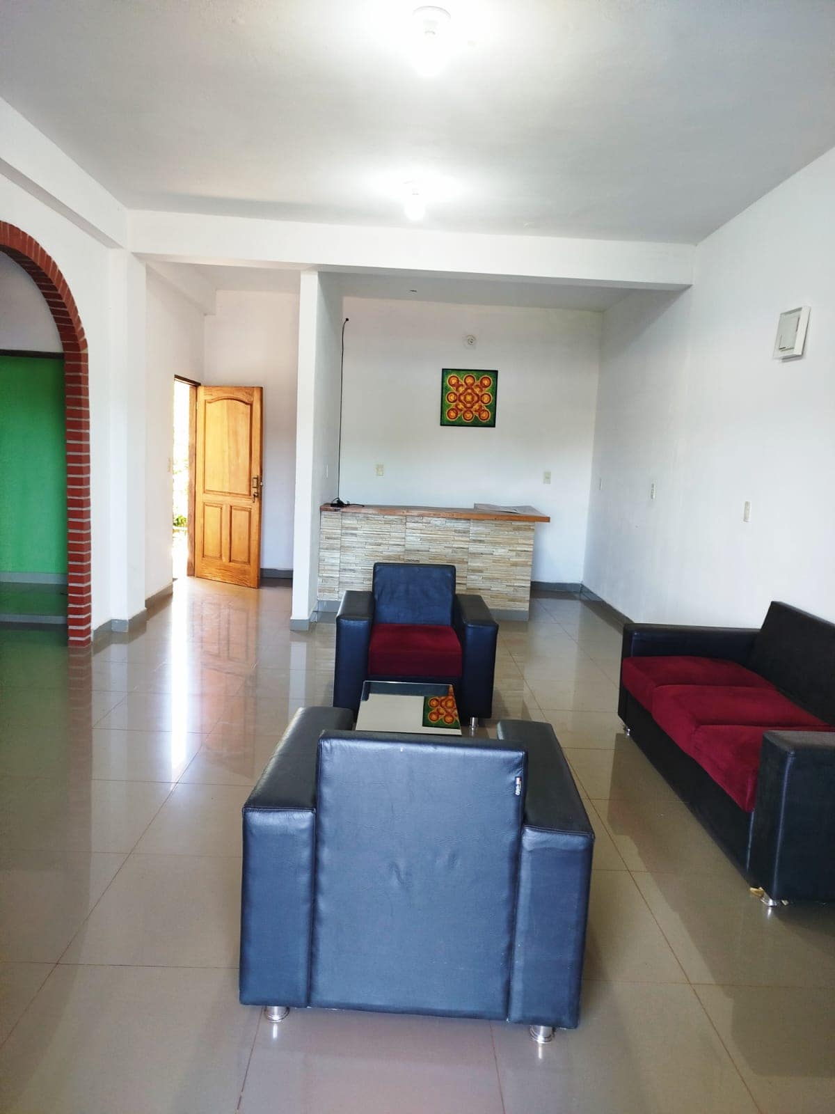 A spacious living area features a mix of seating options, including two black chairs and a red-and-black sofa. A small coffee table is centered among the seating, with a stone-accented bar in the background. Natural light enters through the open doorway, enhancing the room's openness.