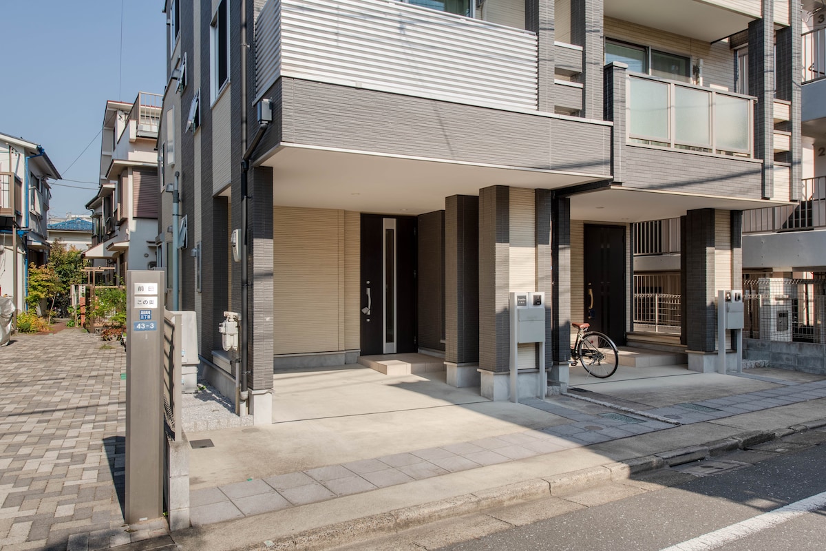 The exterior of a modern apartment building is depicted, featuring a clean entrance with two covered doorways. Paved pathways lead to the doors, with a small bicycle parked close by. The surrounding area includes a well-maintained landscape and a quiet street.
