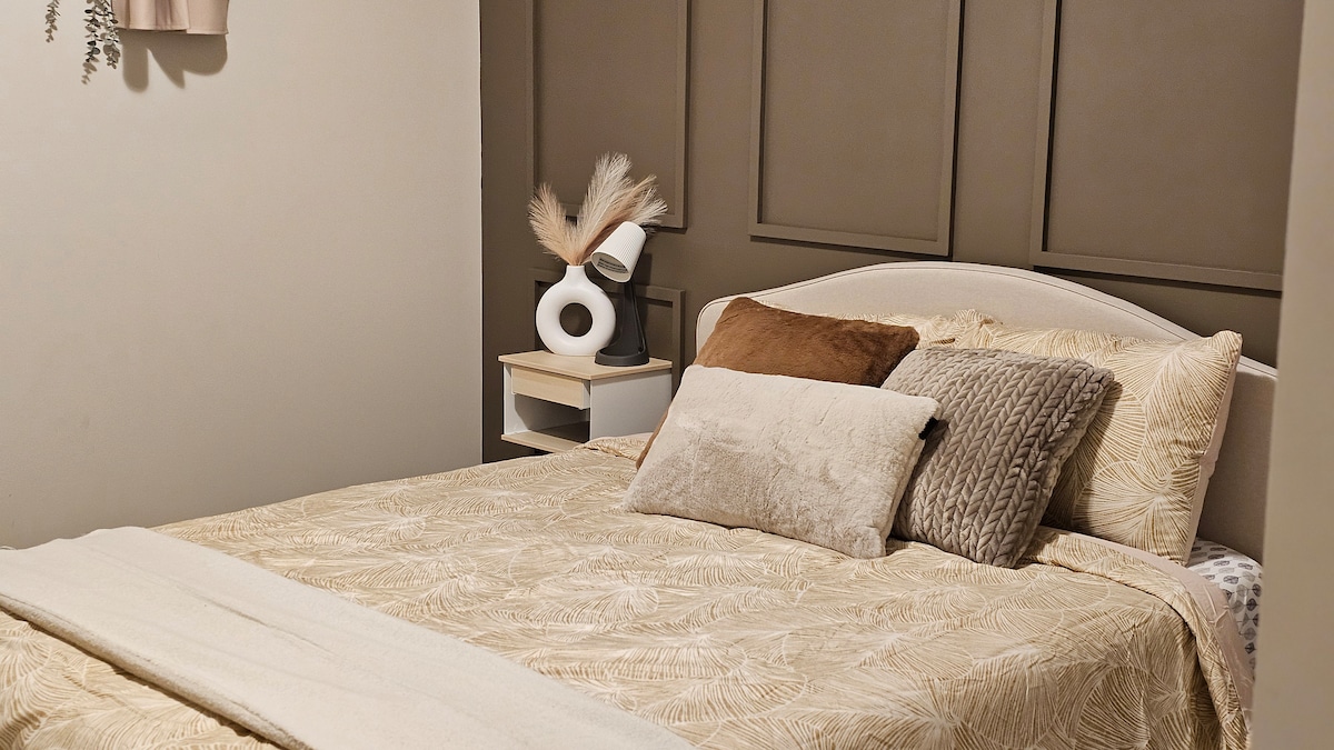 A cozy bedroom features a spacious bed dressed in a patterned blanket and cushioned pillows. A bedside table holds a decorative lamp. Soft earth tones create a calming atmosphere, complemented by wall accents that add depth to the space.