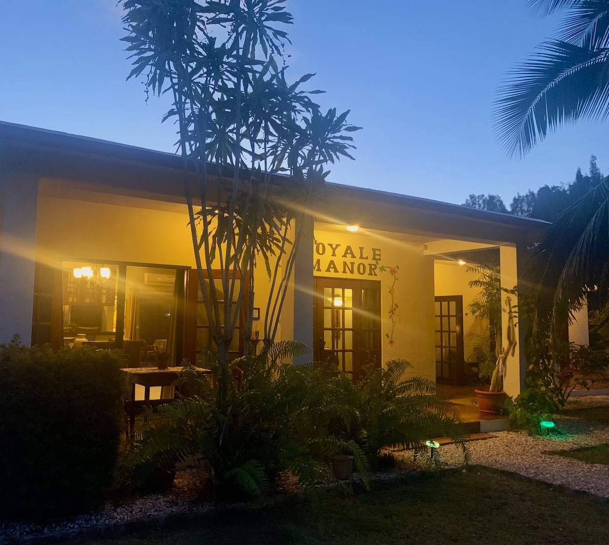 The exterior of the property is softly illuminated, highlighting the entrance marked 'Royale Manor.' Lush greenery, including palm and fern plants, surrounds the entrance, creating a welcoming ambiance. The structure features large glass doors that open to the interior's warmth and inviting atmosphere.