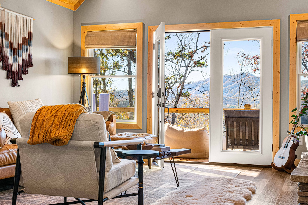 The living area features large glass doors opening to a balcony with mountain views. A comfortable sofa is complemented by a cozy throw and decorative pillows. A small coffee table sits beside a modern floor lamp, while a woven wall hanging adds texture to the space.