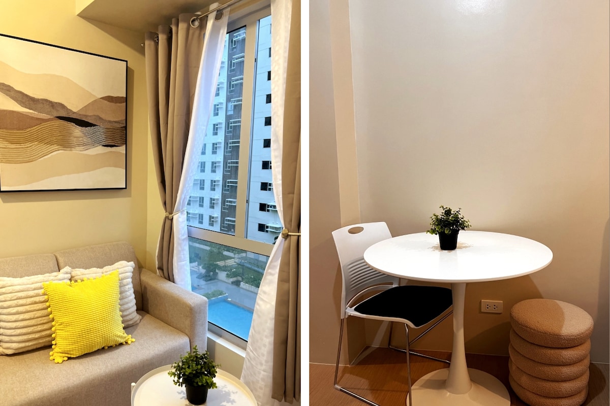 A cozy seating area is highlighted by a light-colored sofa adorned with textured cushions. A piece of abstract art hangs above the sofa, complemented by natural light filtering through the window, offering a view of the outside. A small round table with a plant is also featured.