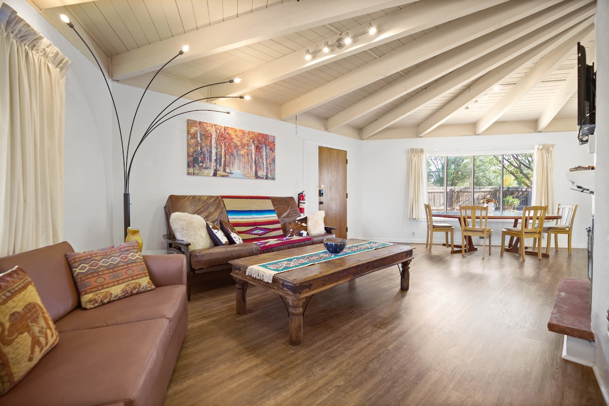 A bright living area features vaulted ceilings supported by wooden beams. Comfortable seating is provided by a brown sofa and a wooden couch, adorned with colorful textiles. An art piece depicting autumn trees hangs above a rustic coffee table, with a dining area visible through large windows.