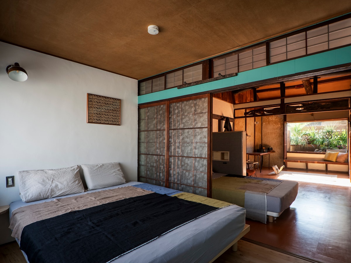 A spacious sleeping area features a large bed with simple bedding in muted tones. Sliding glass doors lead to a living space filled with natural light, showcasing a view of the lush indoor garden beyond. The warm wooden tones of the flooring enhance the inviting ambiance.