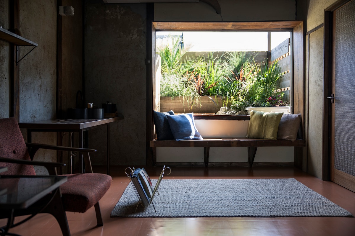 A cozy seating area features a windowed bench with colorful cushions, overlooking a small garden. A wooden table and chairs are arranged nearby, while a woven rug anchors the space. Natural light fills the room, highlighting the warm tones of the decor.