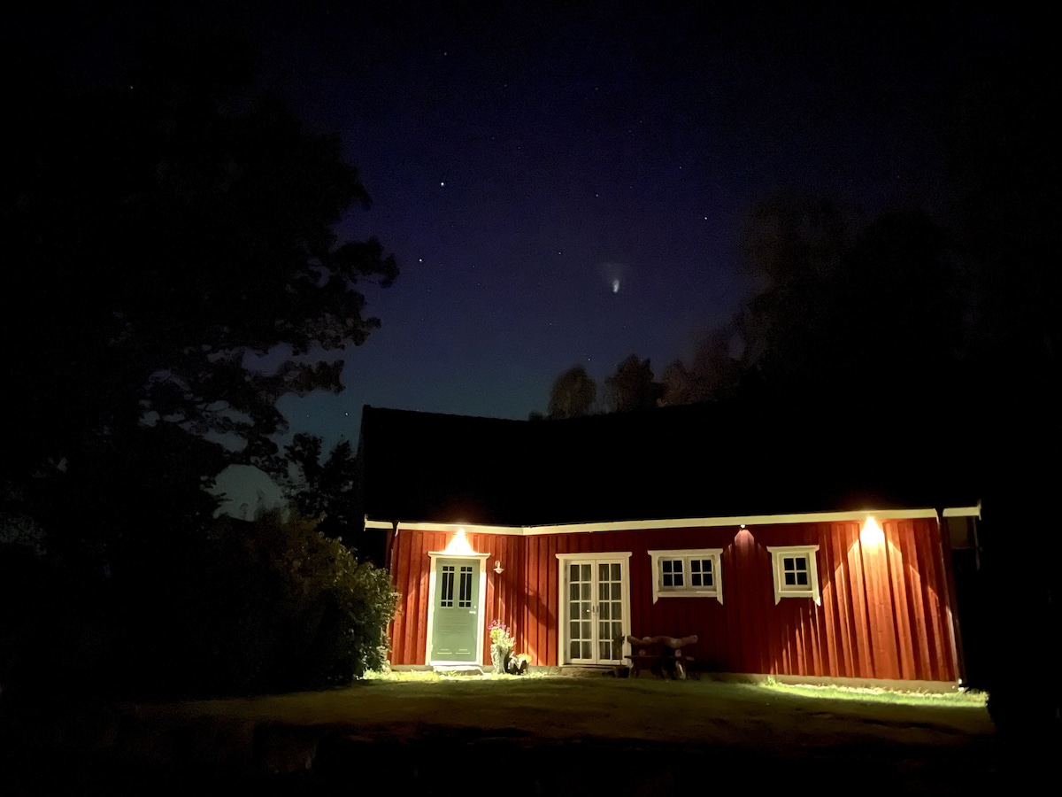 An inviting red wooden cottage is illuminated softly by exterior lighting, showcasing its door and windows. A clear night sky with stars is visible above, providing a serene backdrop. Surrounding trees add to the peaceful ambience of the rural setting.