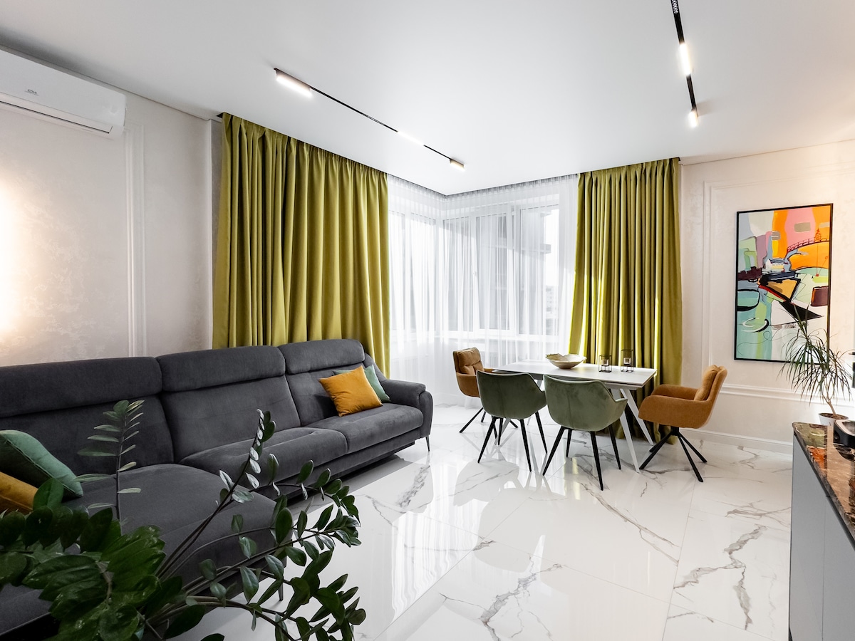 A modern living area is presented, featuring a gray sectional sofa with colorful cushions. A round dining table is surrounded by upholstered chairs. Bright green curtains frame the large window, allowing natural light to brighten the marble flooring and stylish decor.