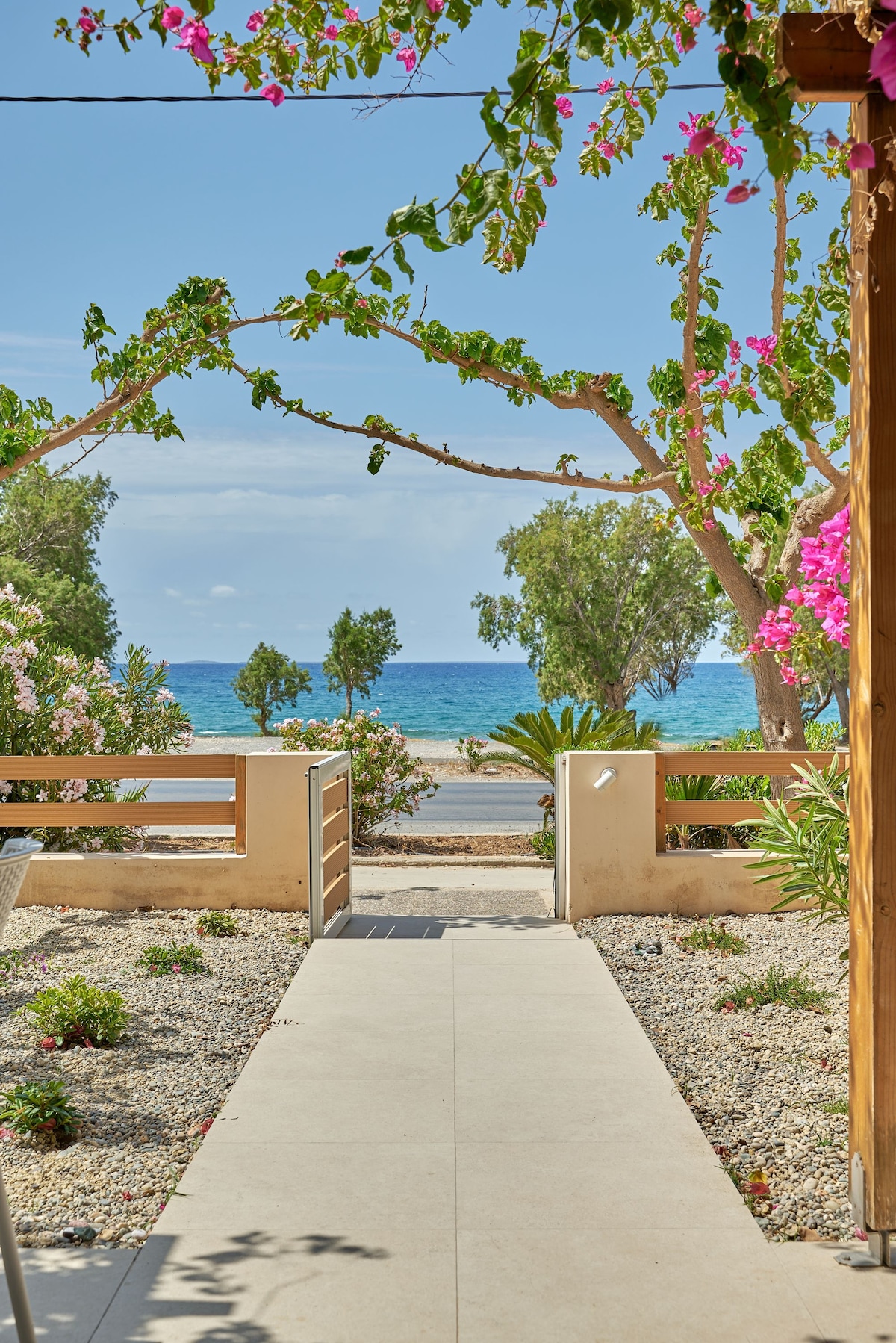 A pathway leads towards the sea, flanked by vibrant greenery and flowering plants. The turquoise water is visible in the distance, with a clear sky overhead. The setting creates a tranquil atmosphere, ideal for enjoying the natural surroundings.