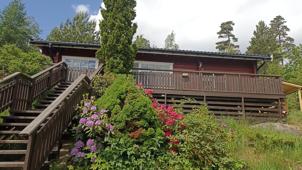 The exterior of a red wooden cabin is showcased, featuring a welcoming front deck with a railing and steps leading up from the garden. Lush green foliage and colorful flowering shrubs surround the stairs, complementing the natural setting.