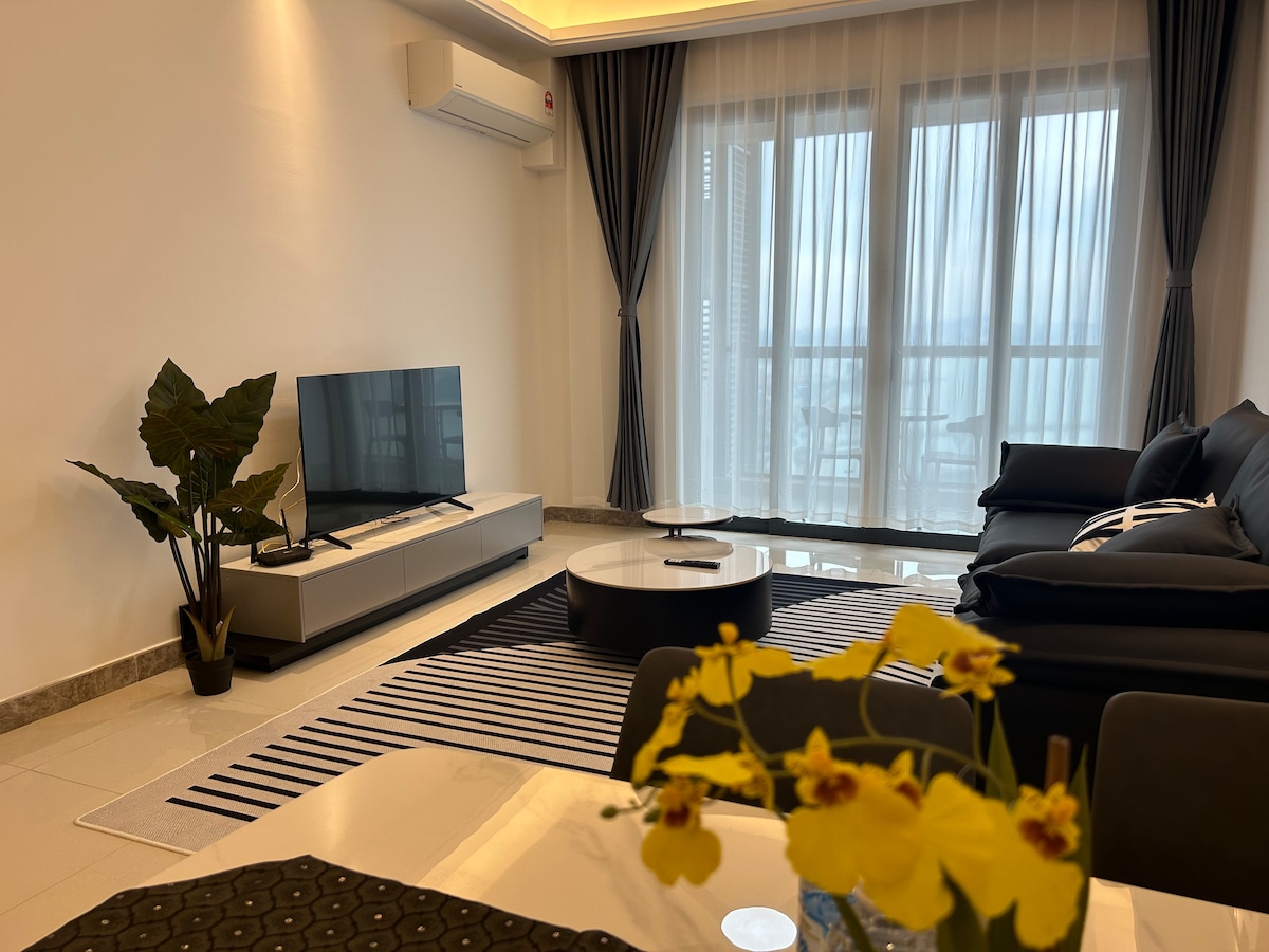 A modern living area is presented with a large flat-screen television positioned on a sleek stand. A dark sectional sofa pairs with a round coffee table on a striped rug. Floor-to-ceiling windows are dressed in sheer curtains, allowing diffused natural light to enter the space.