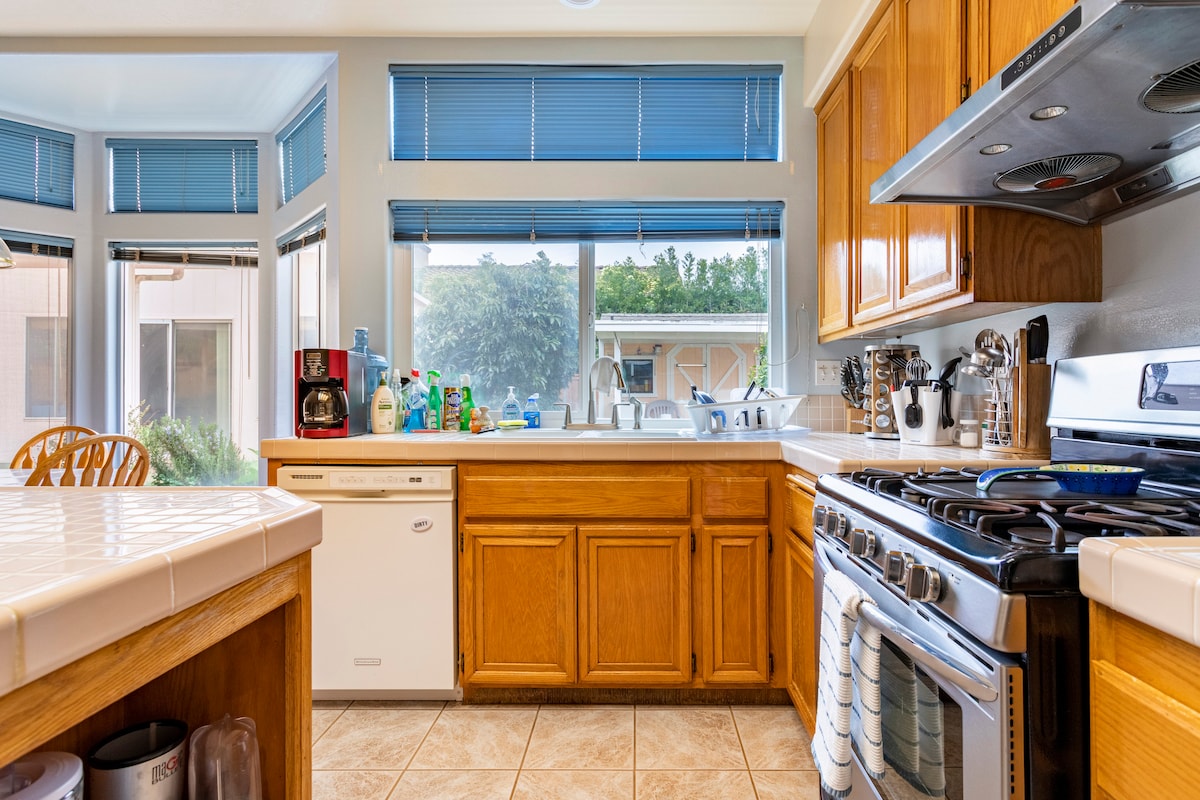 A bright kitchen area features wooden cabinetry and a large window allowing natural light. Appliances include a gas stove, dishwasher, and coffee maker. A tiled counter extends around the space, with dishes and cleaning supplies organized thoughtfully on the countertops.