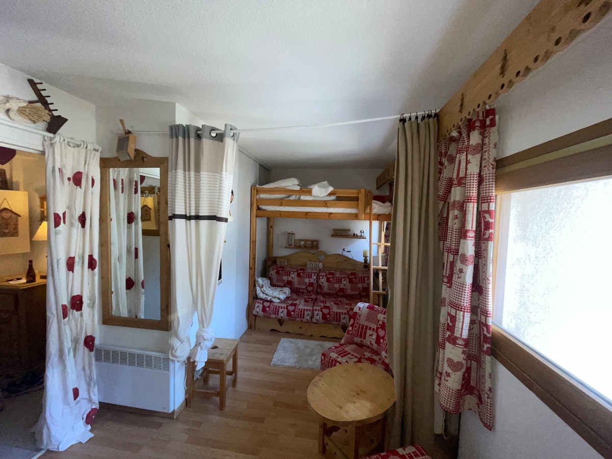 A cozy studio features wooden accents, complemented by red and white patterned furnishings. A bunk bed is visible, along with a comfortable seating area. Natural light filters through the window, creating a welcoming ambiance. A round table is placed nearby, offering a practical dining space.