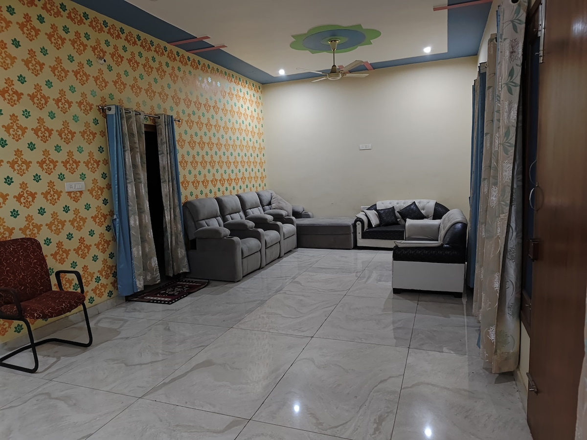 A spacious living area is presented with a mix of comfortable seating, including a large sectional sofa and an accent chair. Brightly colored wallpaper features a floral pattern. Light from ceiling fixtures and natural sources illuminates the glossy tiled floor.