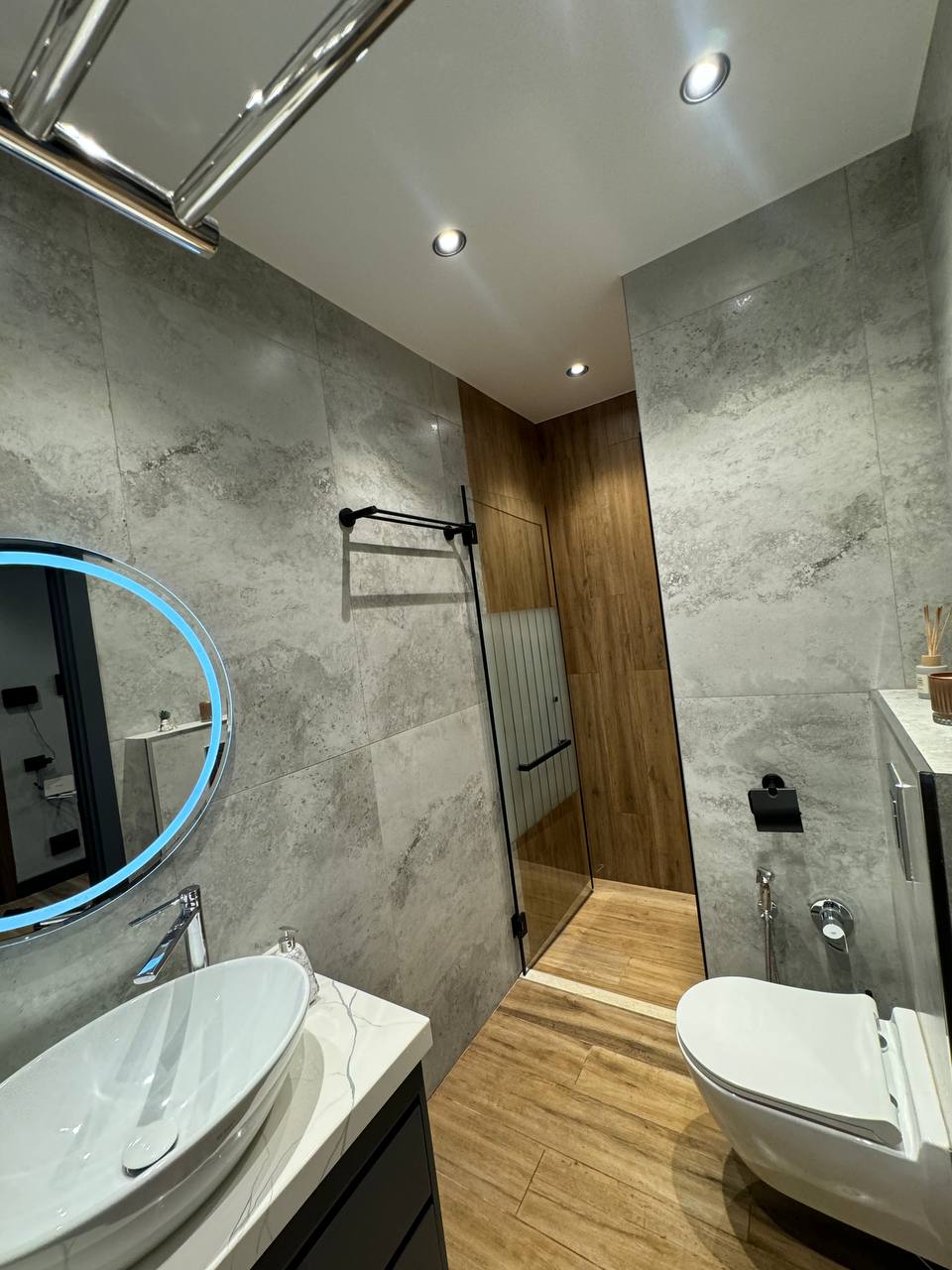 A modern bathroom is presented with sleek gray tiles and warm wooden accents. A circular mirror is mounted above a white sink, with a glass shower stall visible beside it. Lighting is provided by recessed fixtures, creating a well-lit, inviting space.