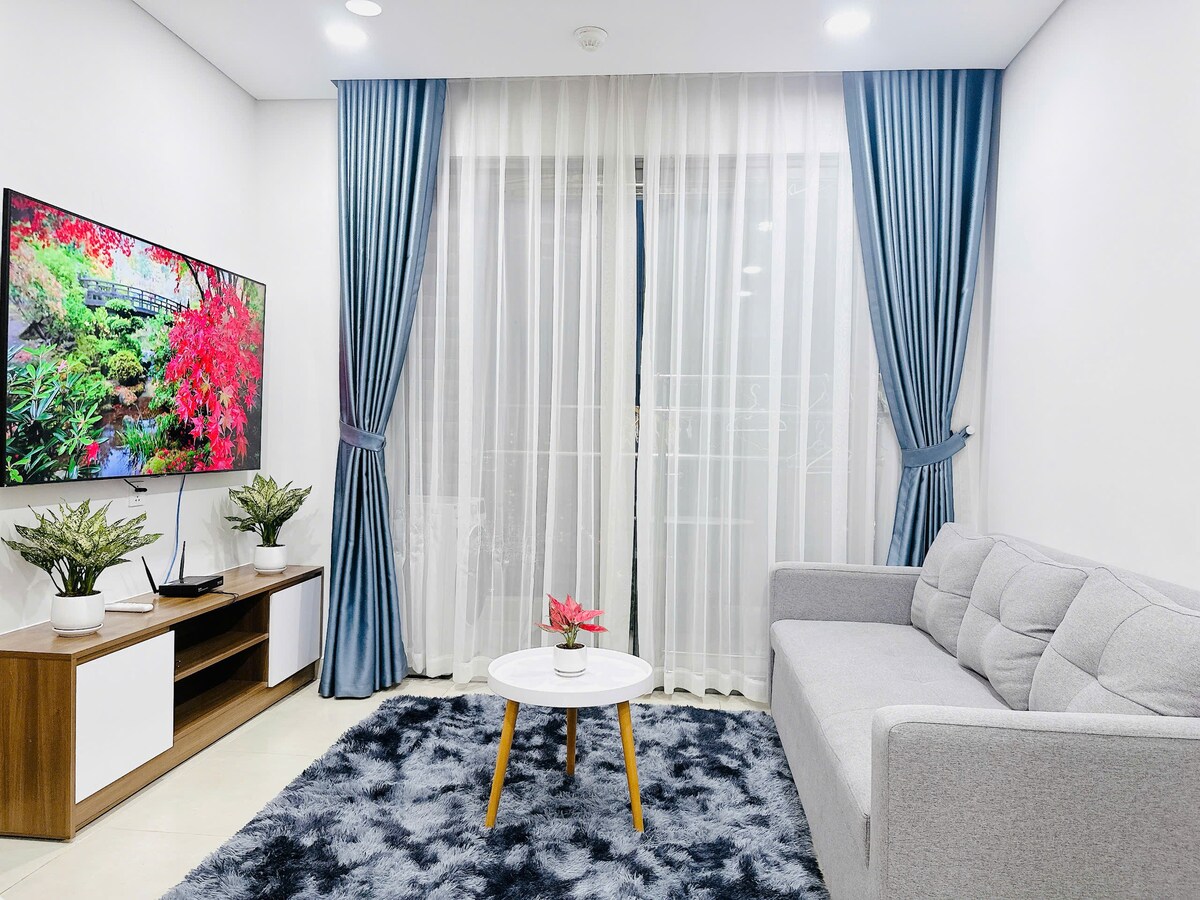 A bright living area features a modern sofa arranged beside a low coffee table. A large television is mounted on the wall opposite. Light-colored curtains frame the windows, allowing natural light to fill the space, which is enhanced by decorative indoor plants.