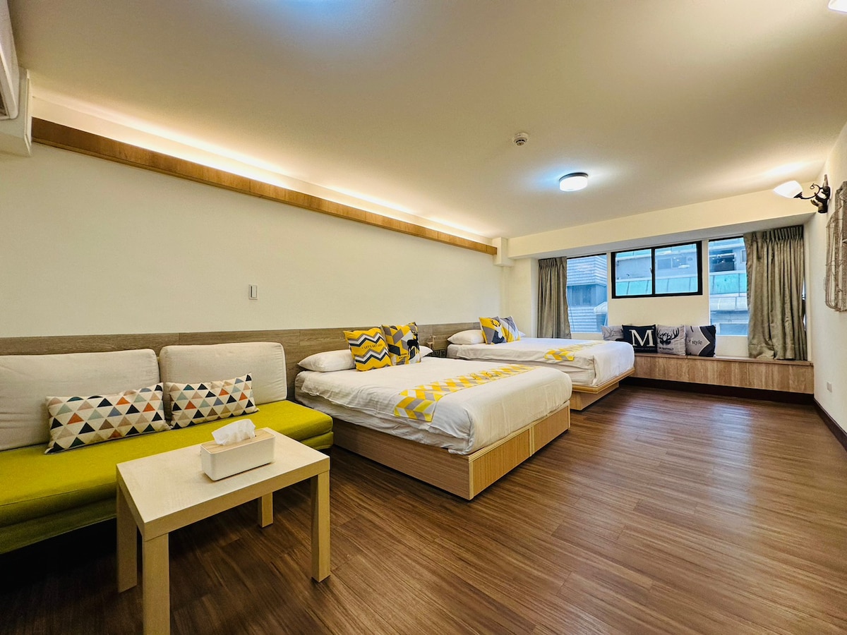 A spacious room features two standard double beds with minimalist bedding and patterned throw pillows. A cozy sofa and a small coffee table are positioned near large windows adorned with light curtains, allowing natural light to fill the space.