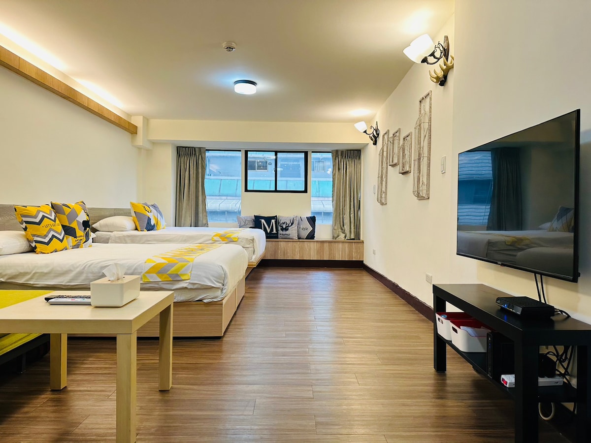 An open space is presented with two spacious beds, accented by decorative cushions in a chevron pattern. A flat-screen TV is positioned on a media console, and large windows provide natural light. A seating area by the window features a comfortable bench.