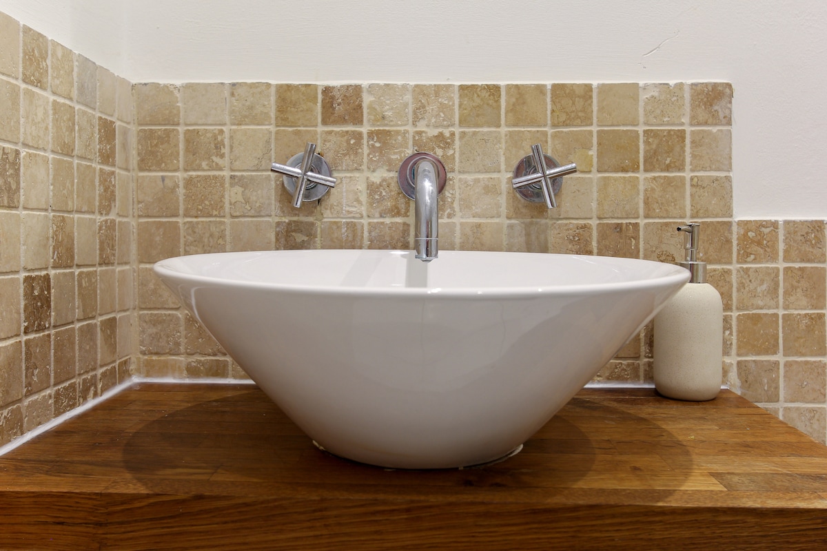 A modern bathroom sink is displayed, featuring a sleek, bowl-shaped design positioned on a polished wooden countertop. The wall behind is adorned with square tiles in warm beige tones, complemented by contemporary faucet fixtures. A soap dispenser stands next to the sink, enhancing the minimalistic aesthetic.