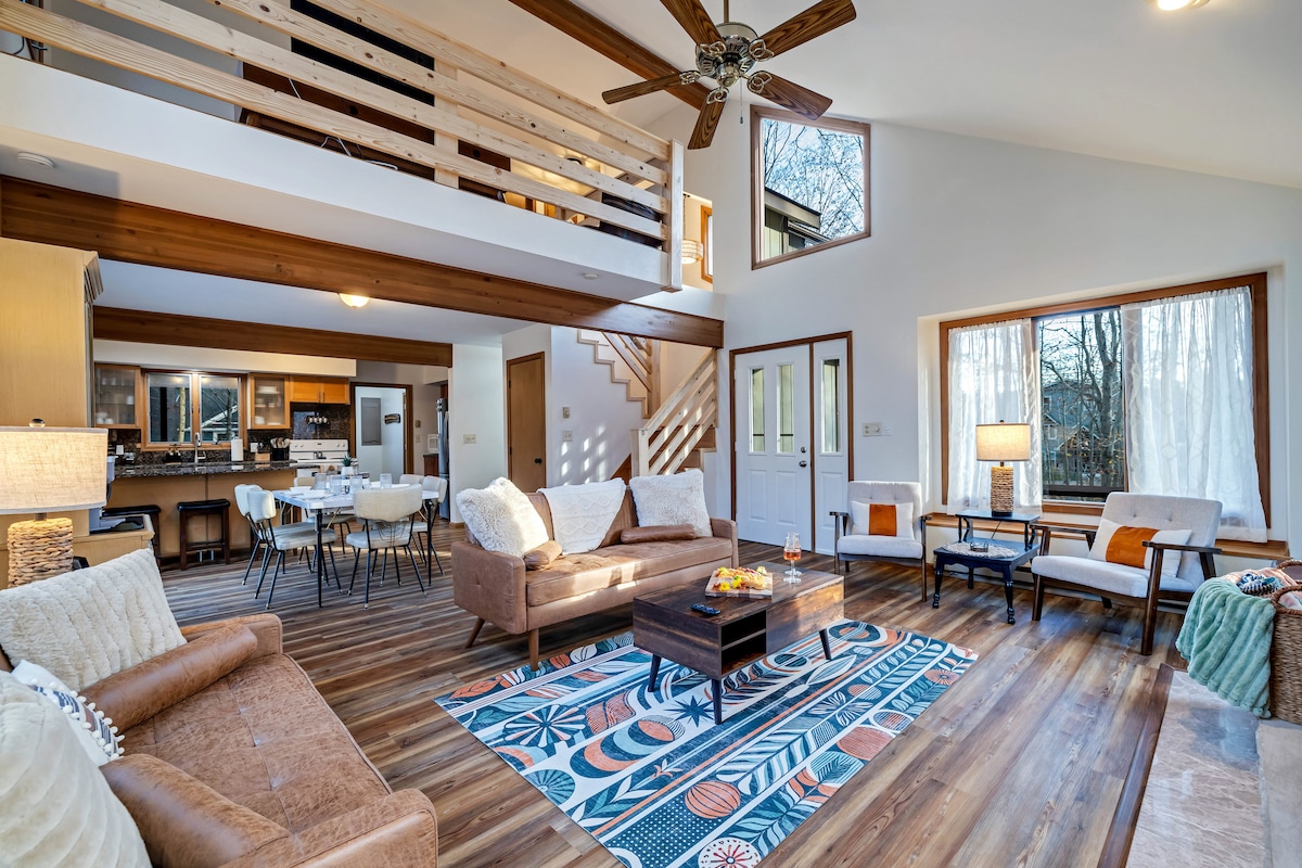 A spacious living area features large windows allowing natural light to fill the room. Comfortable couches and armchairs encircle a coffee table on a colorful area rug. The open floor plan connects to a dining area and kitchen, with wooden beams adding to the inviting atmosphere.