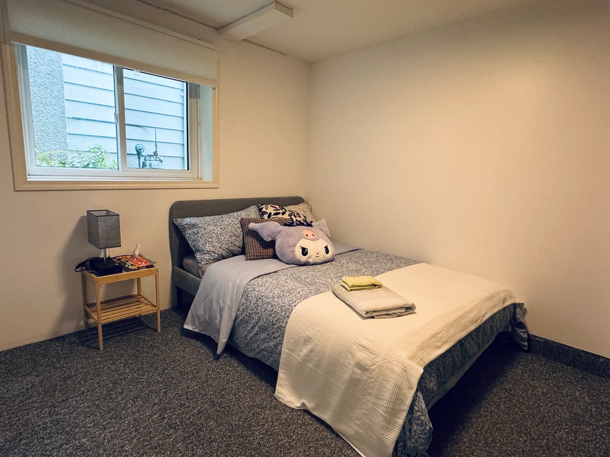 A cozy bedroom is presented with a spacious bed, adorned with pastel bedding and a decorative pillow. A small bedside table holds a lamp, while a window allows natural light to brighten the room, revealing a simple and clean decor.