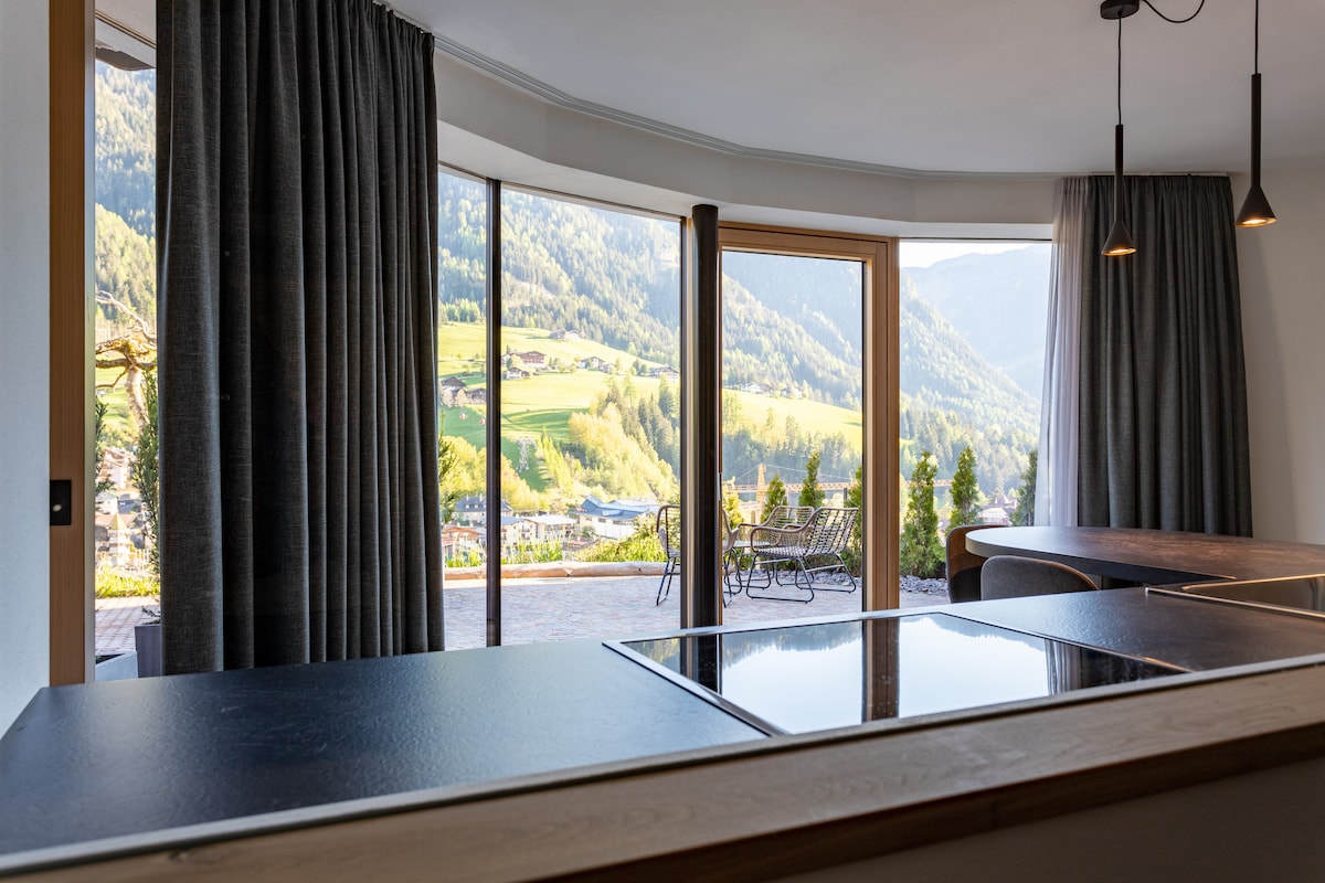A spacious kitchen area is visible, featuring a modern black countertop and a sleek cooktop. Large glass windows provide a panoramic view of the surrounding mountains and valleys, while elegant curtains frame the scene, allowing natural light to enter.