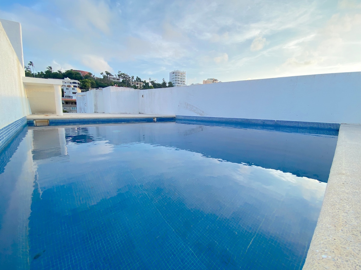 A shared pool is displayed with tranquil blue water, reflecting the sky. Surrounded by a simple white wall, the area offers an open and airy feeling, ideal for relaxation. The skyline and distant hills are partially visible in the background.