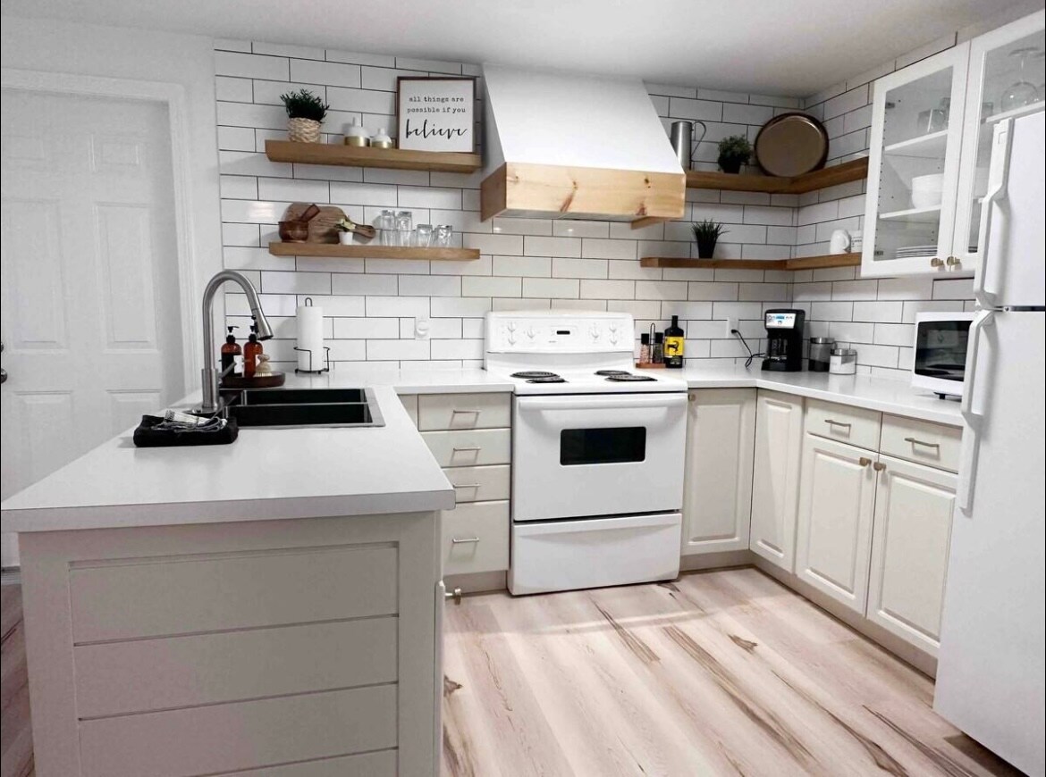 A modern kitchen features white cabinetry and open shelving, displaying glassware and plants. A black sink is situated beside a white stove, while a microwave and coffee maker are positioned on the counter. Natural light illuminates the wood-look flooring.