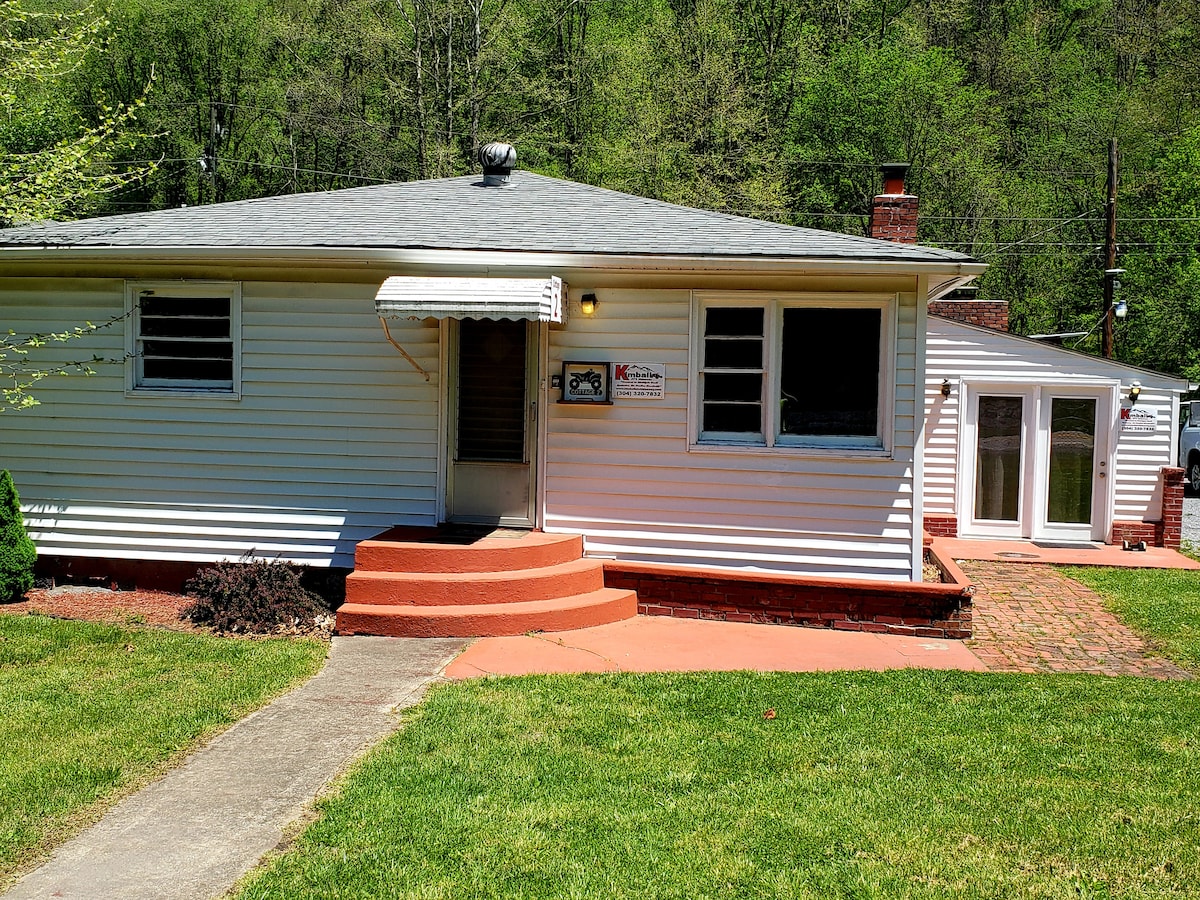 The cottage exterior is characterized by a white façade and a gentle sloped roof. Three windows are visible in the front, allowing natural light. A series of steps lead up to the front door, which is covered by an awning. A green lawn surrounds the entrance.