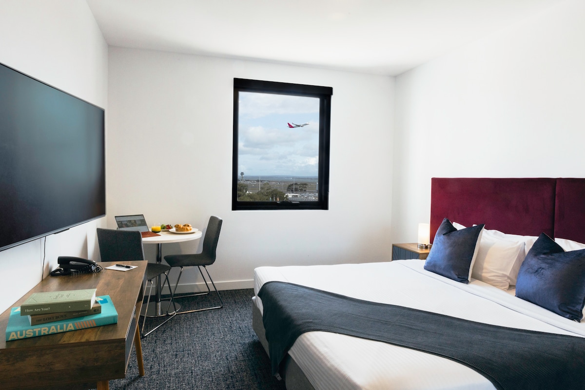 A bright studio apartment features a spacious bed with dark blue bedding and plush pillows. A window offers a view of the sky and passing airplane. A small dining table is set for two with refreshments, while a television sits mounted on the wall.
