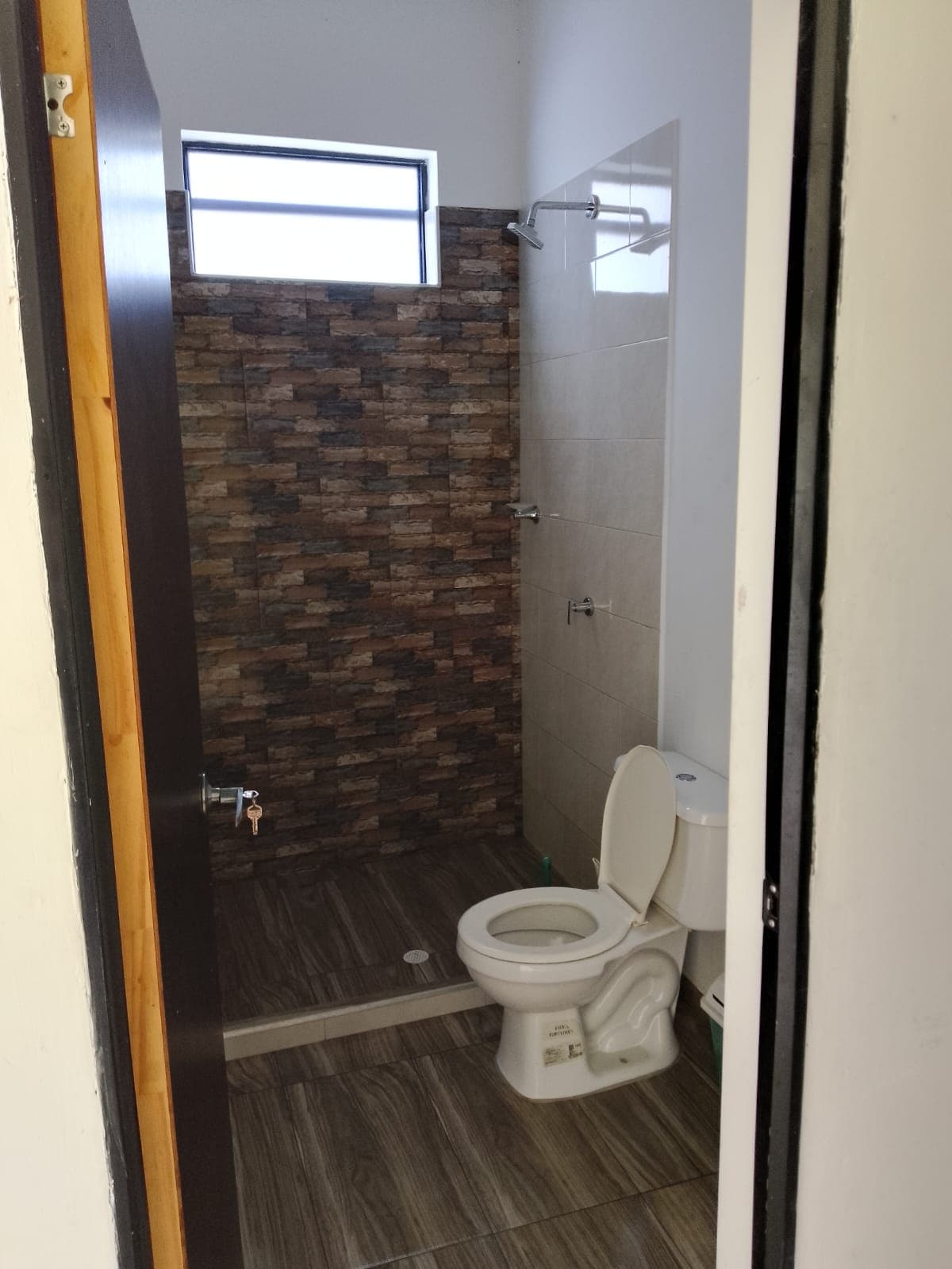 The bathroom features a walk-in shower enclosed by textured stone tiles. A modern toilet is positioned next to the shower, and a sleek sink is visible in the corner. Natural light enters through a small window, illuminating the neutral-toned tiles on the floor.