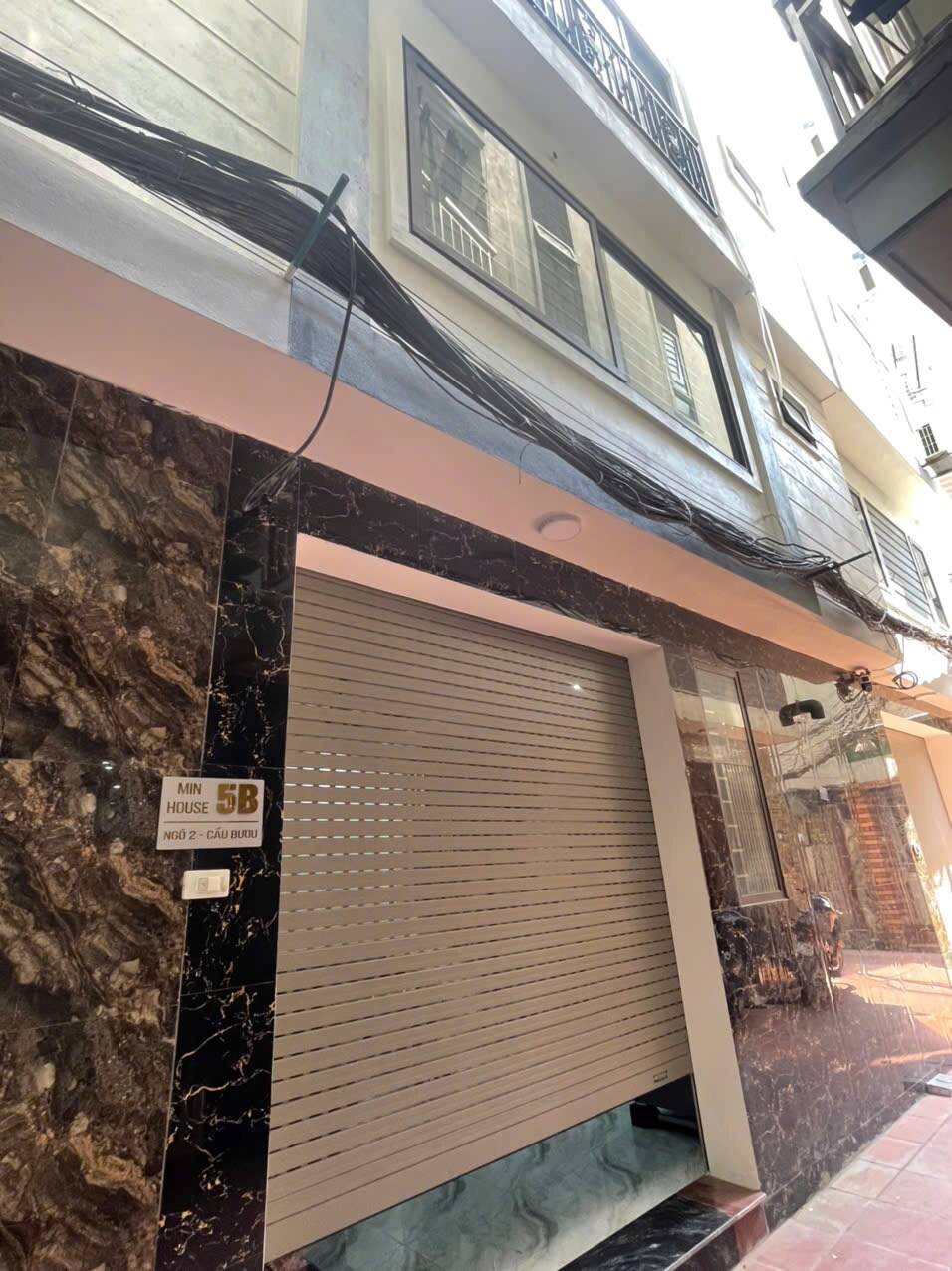 The exterior of a modern building features a sleek marble facade and a garage door with horizontal slats. Above, large windows allow natural light to enter, while electrical wiring runs along the wall. The surrounding area includes narrow passageways between neighboring structures.