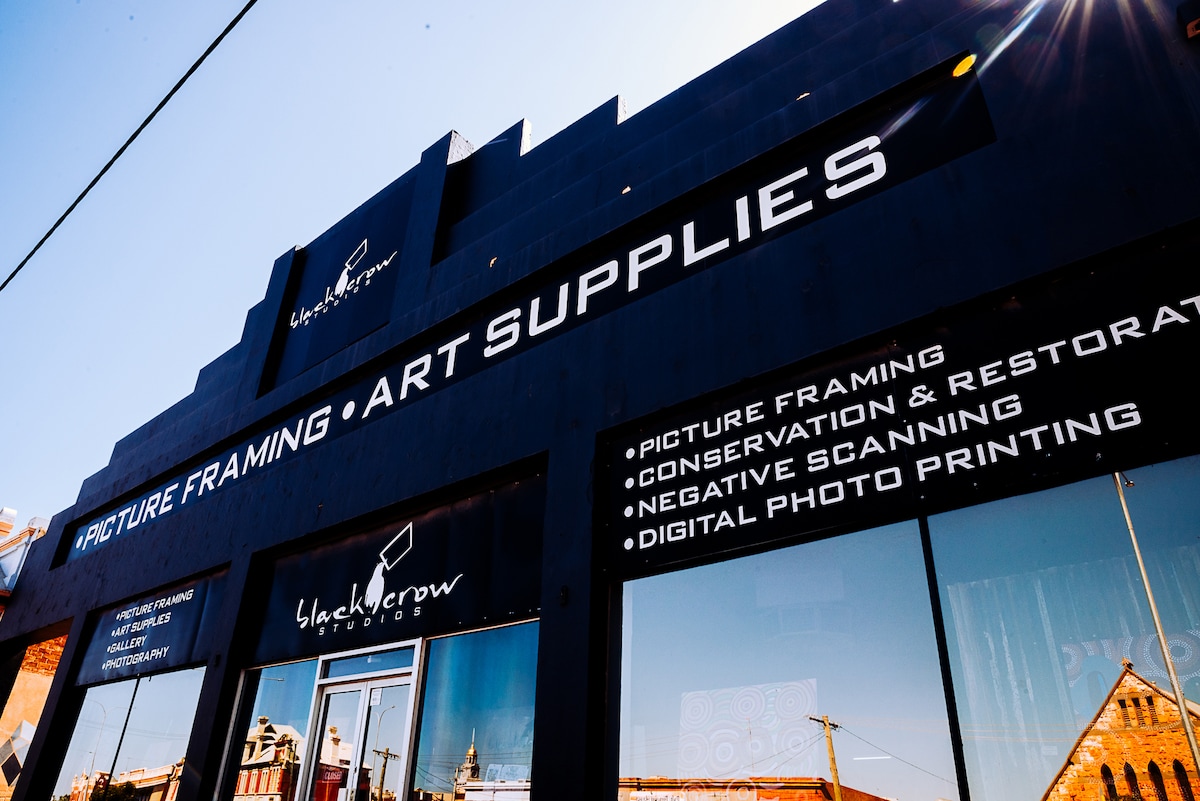 The exterior of a black building features large windows, showcasing the business’s focus on picture framing and art supplies. Bold letters proclaim services such as conservation, restoration, and digital photo printing. A clear blue sky provides a contrasting backdrop.