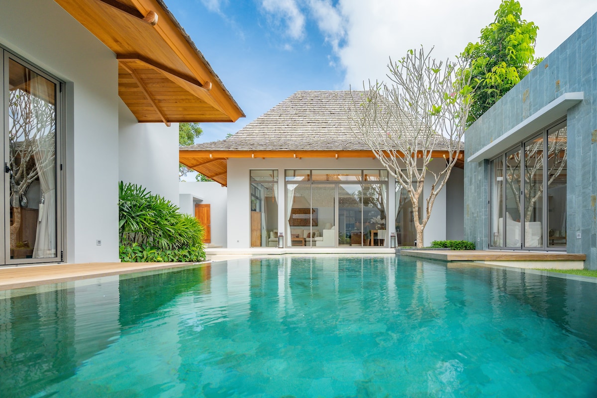 A private swimming pool reflects the clear sky and lush greenery. Surrounding the pool are modern architectural elements, including large glass doors that create an open connection to the interior spaces. The area is framed by tropical trees and a wooden overhang, enhancing the tranquil setting.