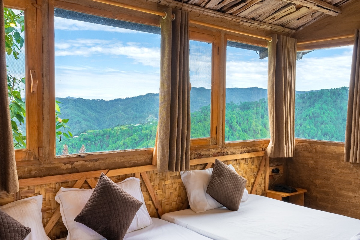 Large windows frame scenic views of the surrounding mountains and greenery, allowing natural light to fill the inviting sleeping area. Two spacious beds are placed against textured walls, accompanied by pillows featuring neutral tones. Soft curtains gently complement the rustic decor.